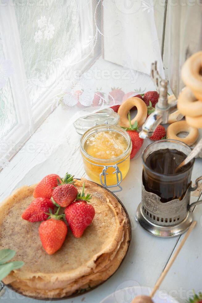 ainda vida dentro a russo tradição para maslenitsa panquecas com querida e morangos, chá dentro lapidado vidro com bagels foto