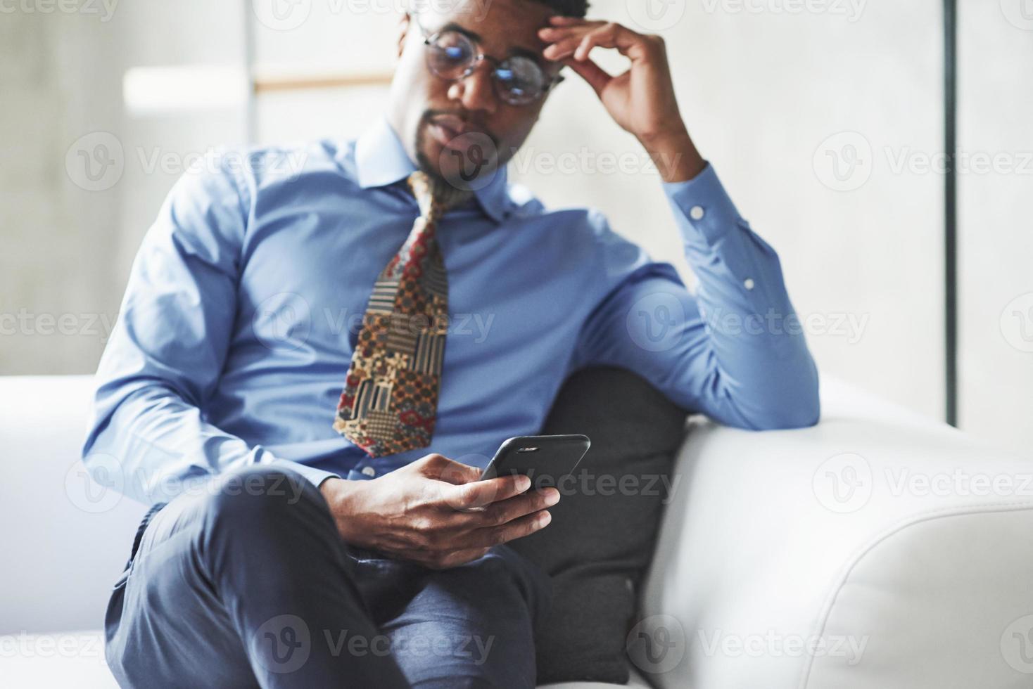 habing preocupações sobre o negócio. foto de jovem negro elegante de terno e óculos segurando e olhando para o telefone enquanto está sentado no sofá