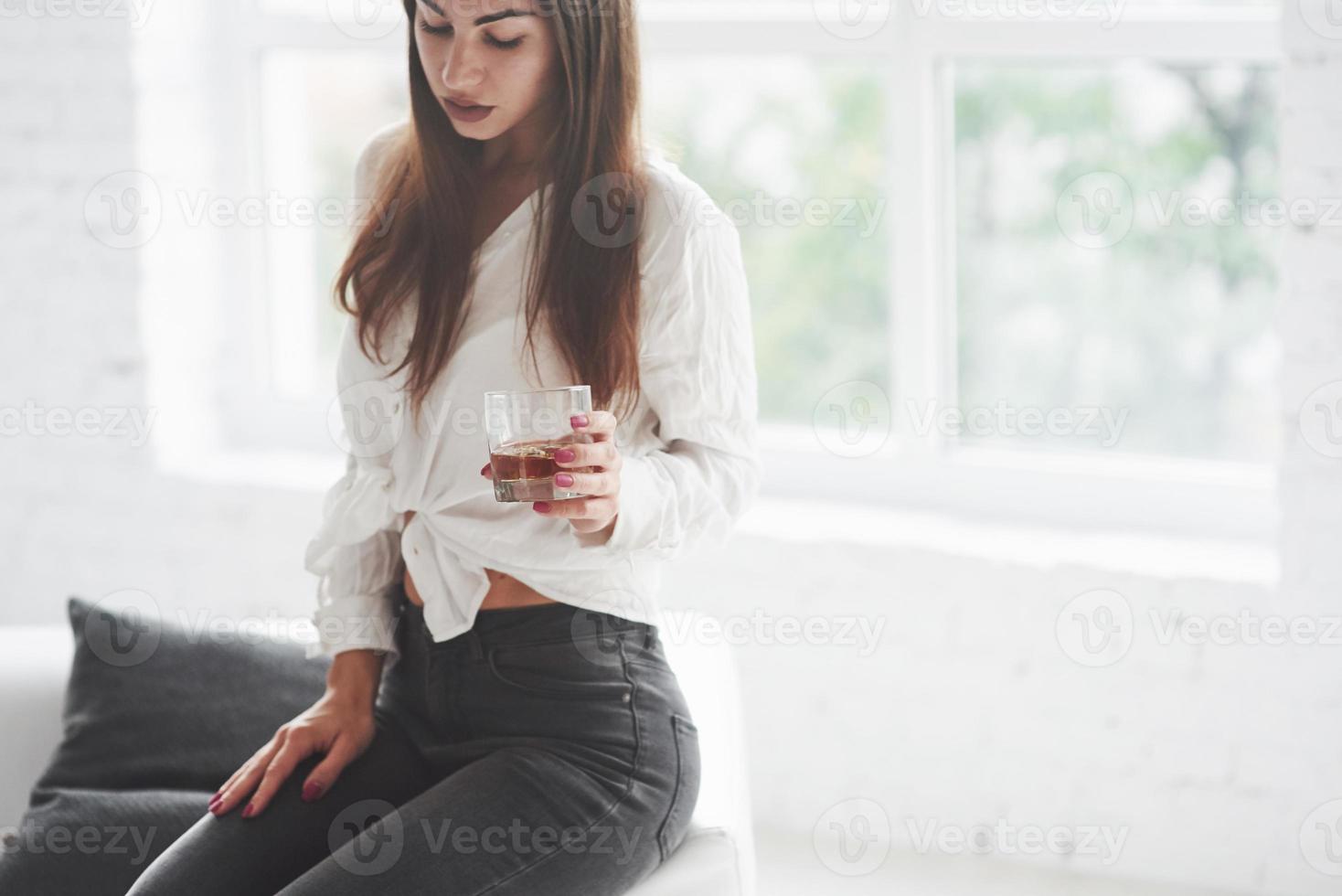 feche a foto de uma linda jovem em jeans preto sentado e segurando um copo com uísque