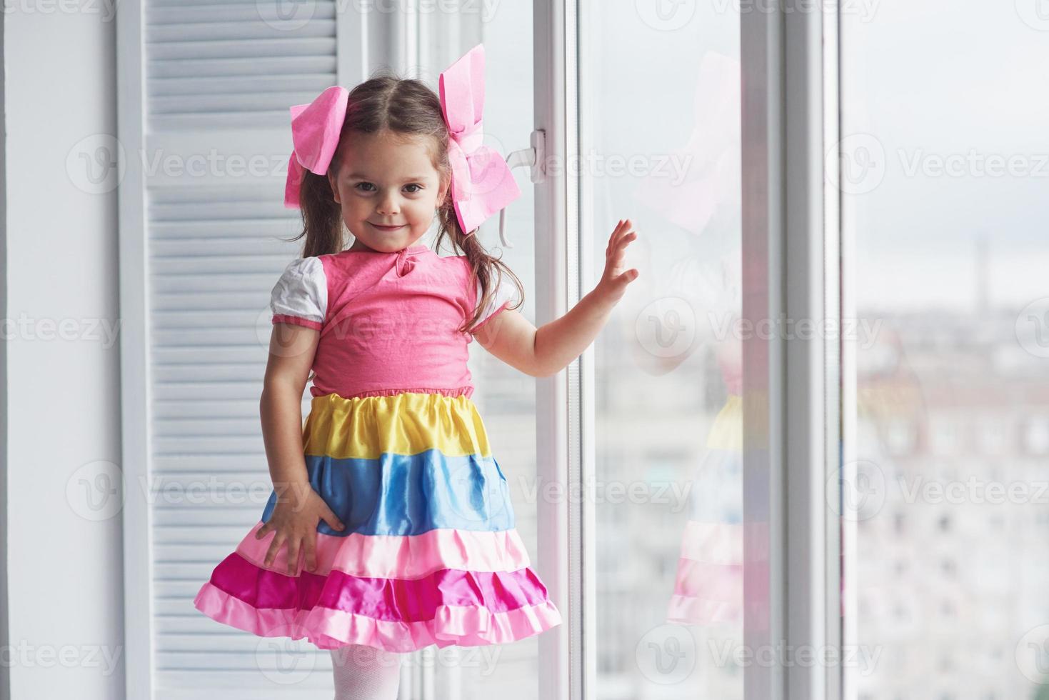 garota de vestido colorido tocando o vidro da janela e olhando para a câmera foto
