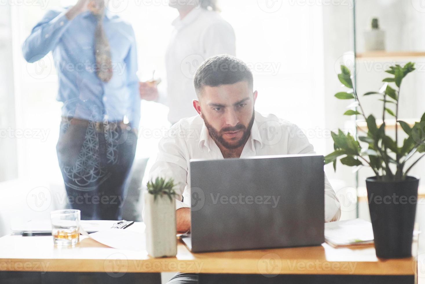 homem barbudo trabalhando com laptop na sala de escritório com dois funcionários no fundo foto