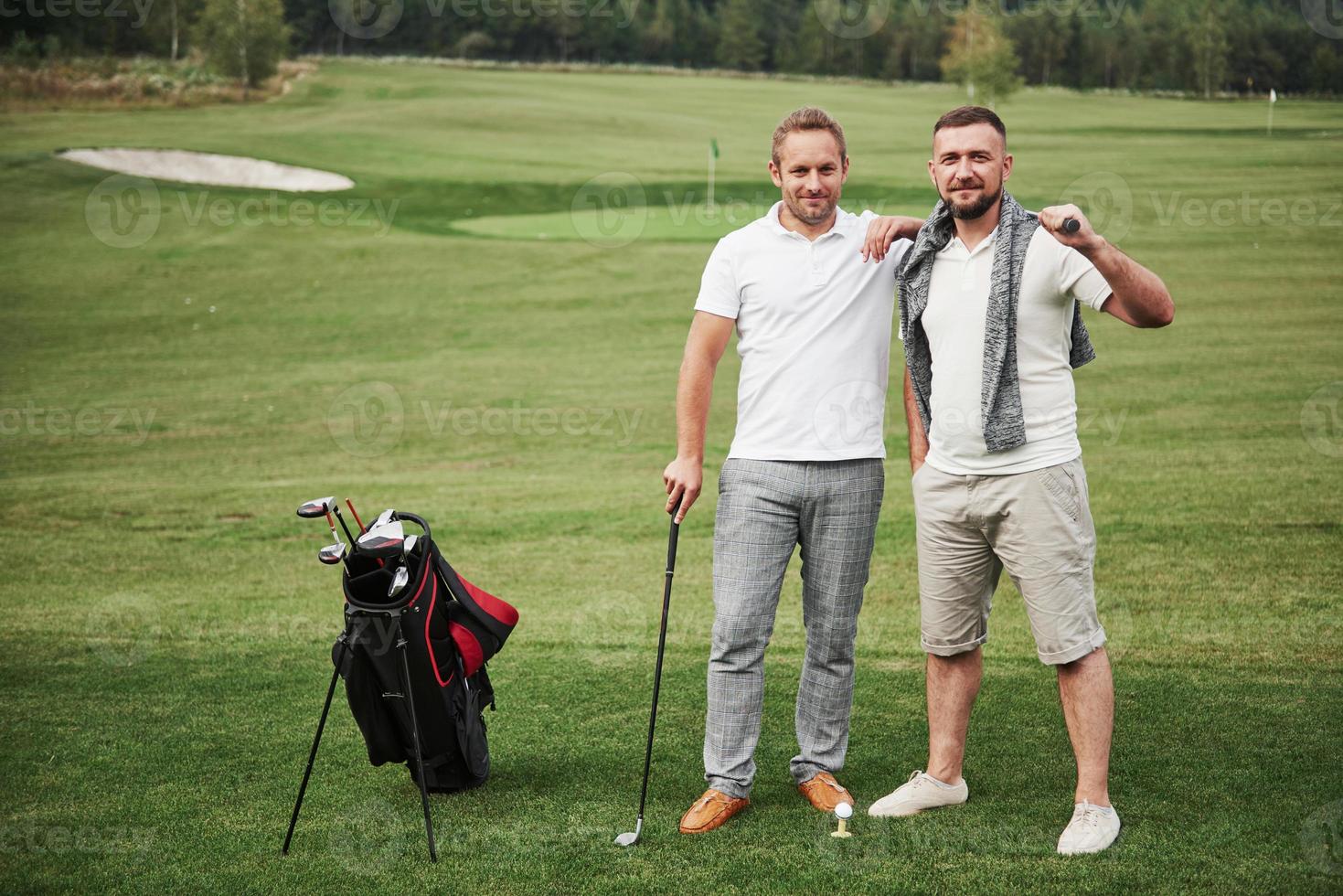 dois homens elegantes segurando bolsas com tacos e caminhando no campo de golfe foto