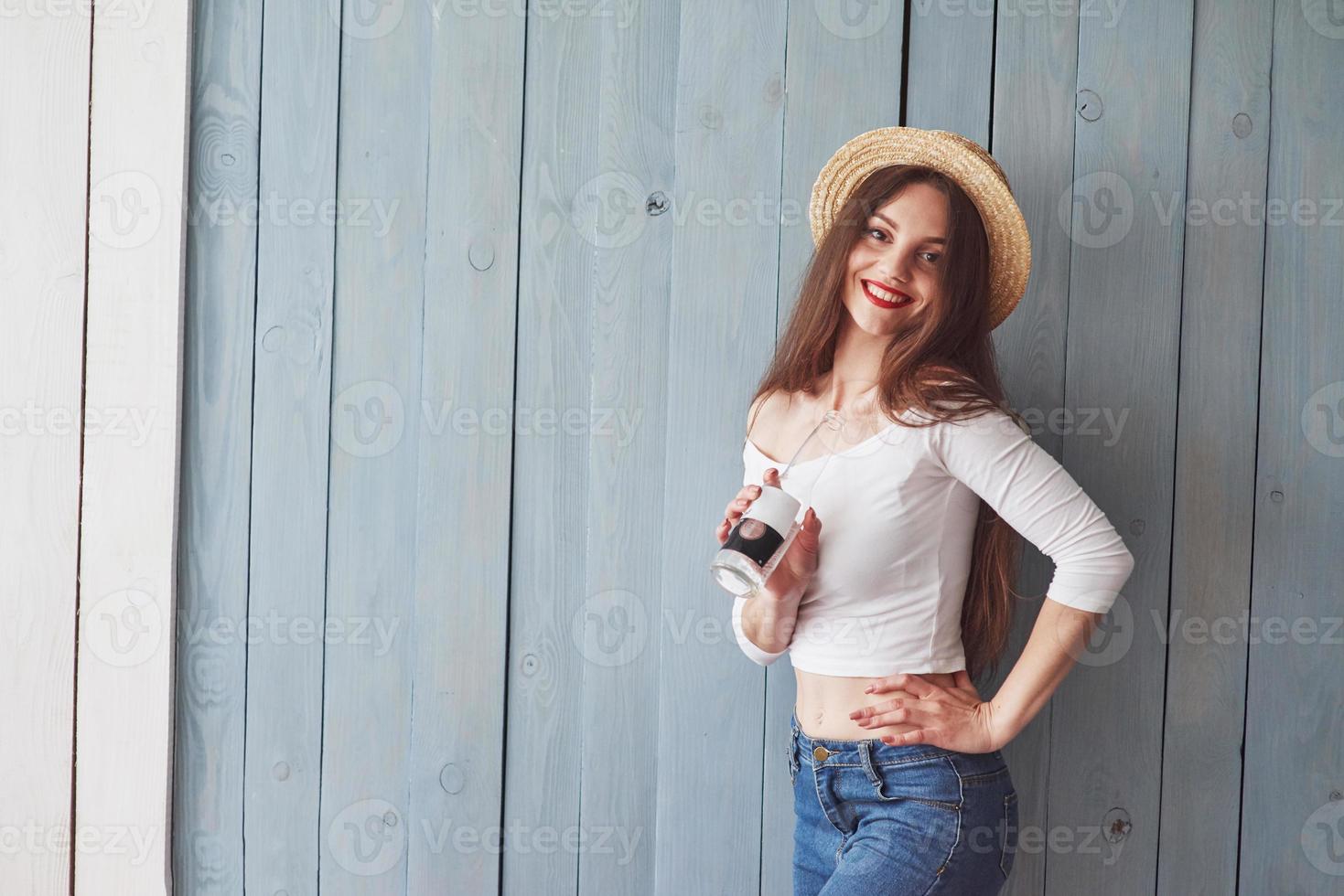 retrato de uma linda jovem com chapéu na cabeça e garrafa na mão em pé contra a parede de madeira foto