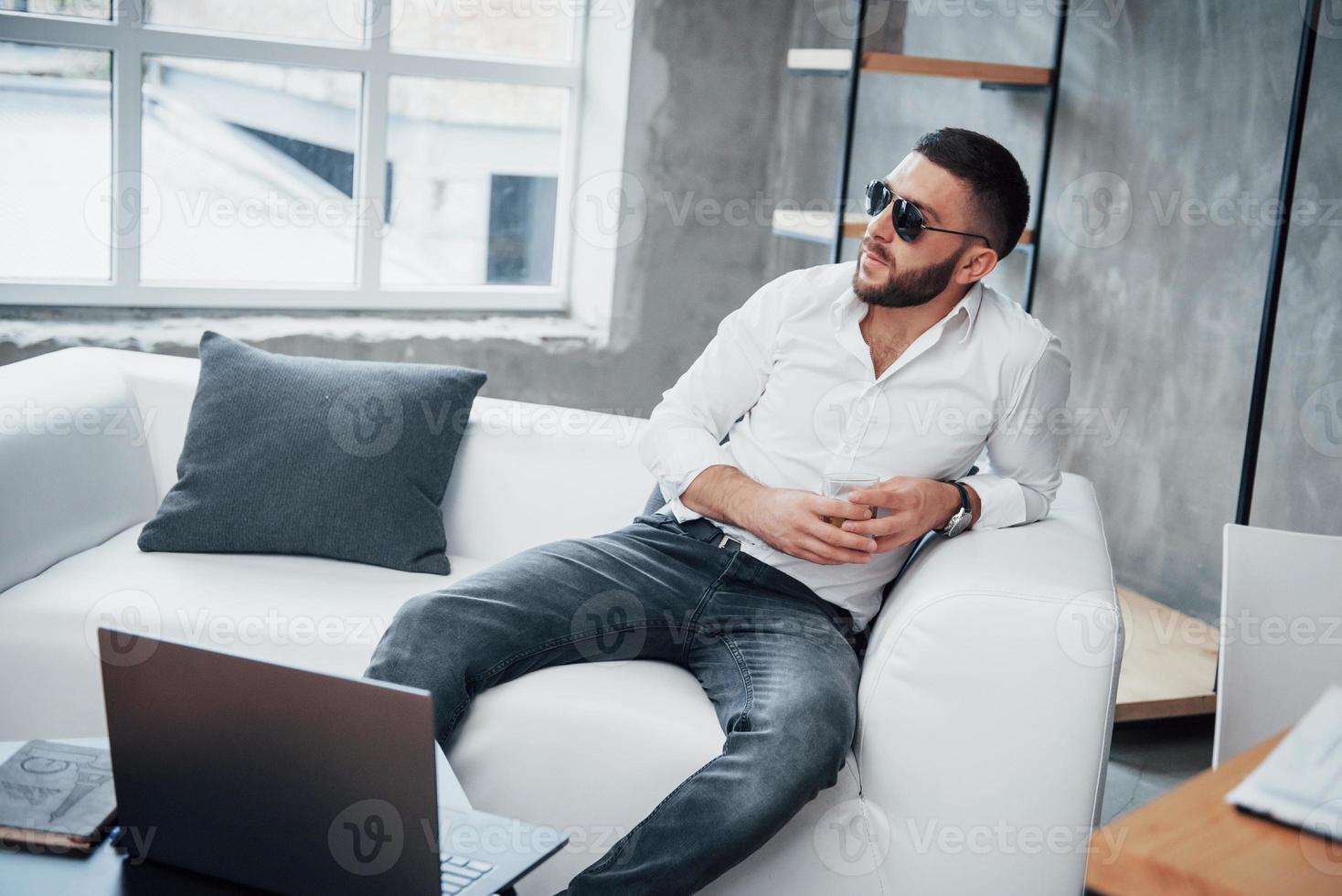 laptop na frente. jovem homem de cabelo curto com óculos escuros sentado no sofá do escritório foto