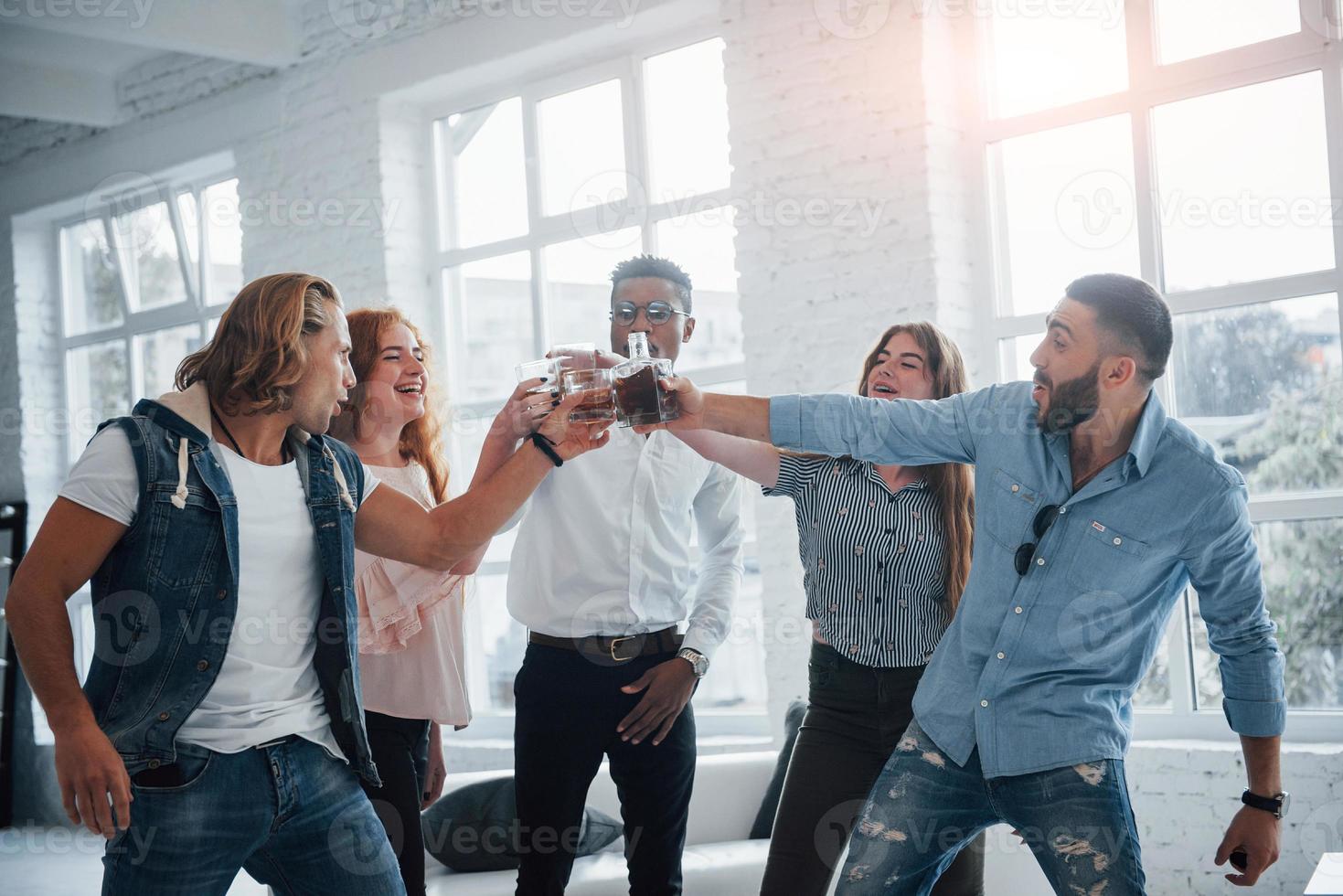 festejando e comemorando. jovens se divertindo no escritório. sorrindo e bebendo com janelas ao fundo foto