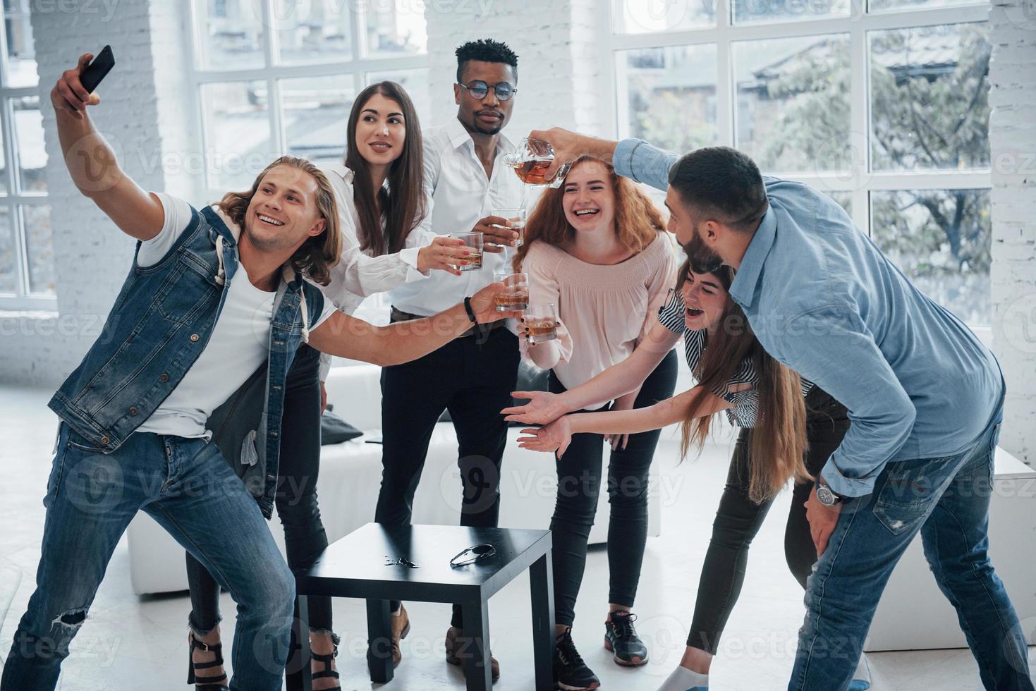 hora de selfie, olhe para o telefone. jovens amigos alegres se divertindo e bebendo no interior branco foto