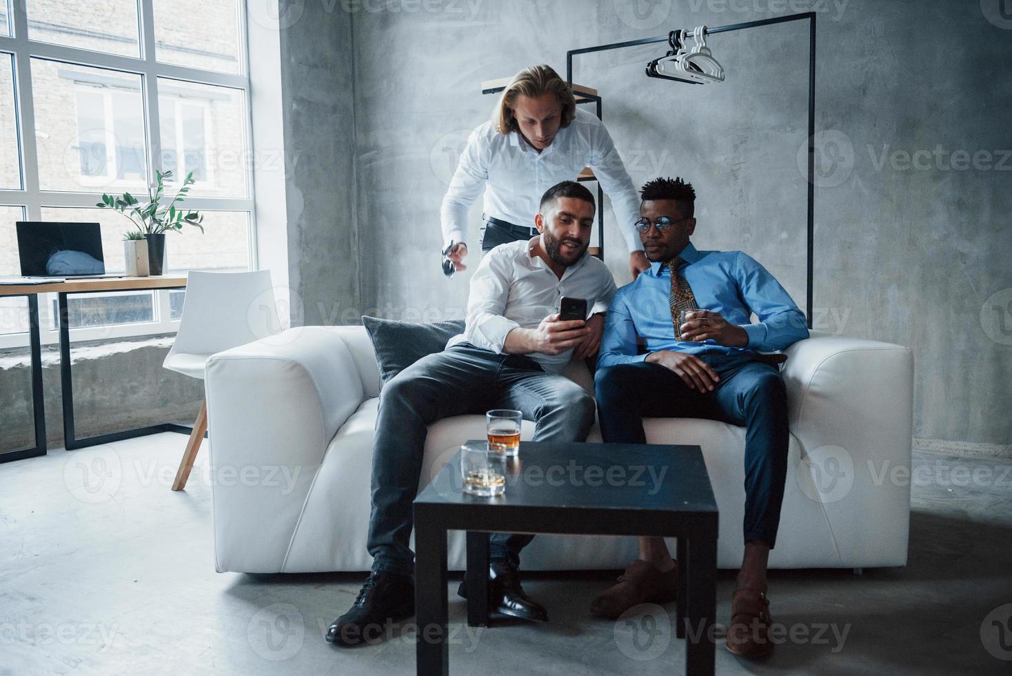 homem com cabelo curto, mostrando informações interessantes no smartphone. grupo de funcionários multirraciais em roupas formais conversando sobre tarefas e planos foto