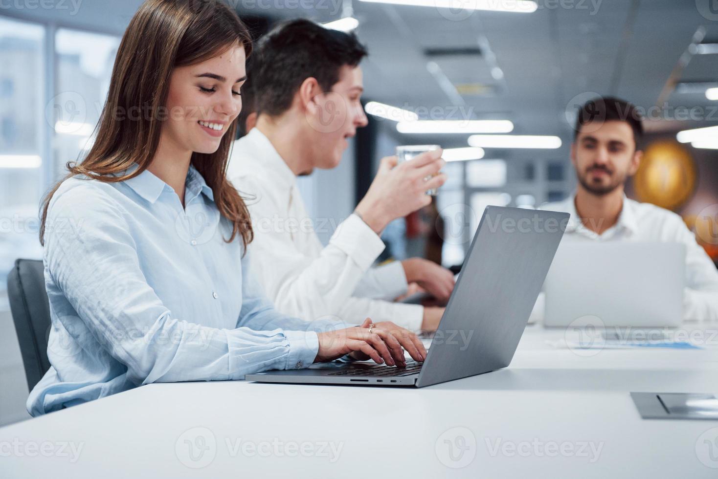 mulher digita um texto enquanto o cara atrás bebe de um copo de prata. vista lateral da garota trabalhando no laptop prateado no escritório e sorrindo foto