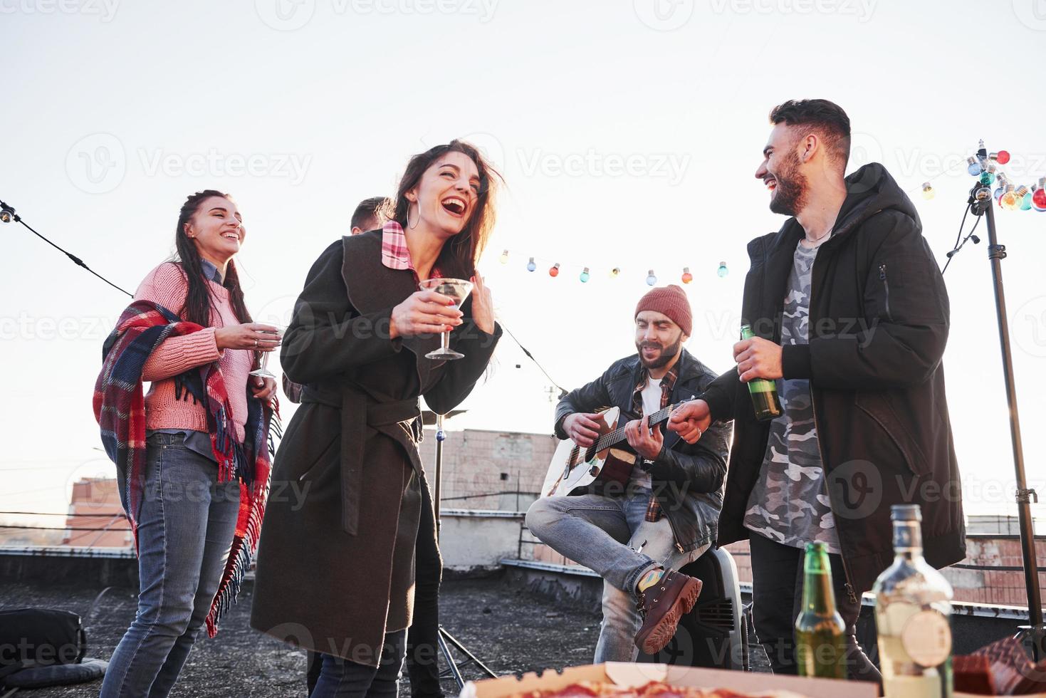 cara canta uma música engraçada. jovens alegres, sorrindo e bebendo no telhado. pizza e álcool na mesa. guitarrista foto