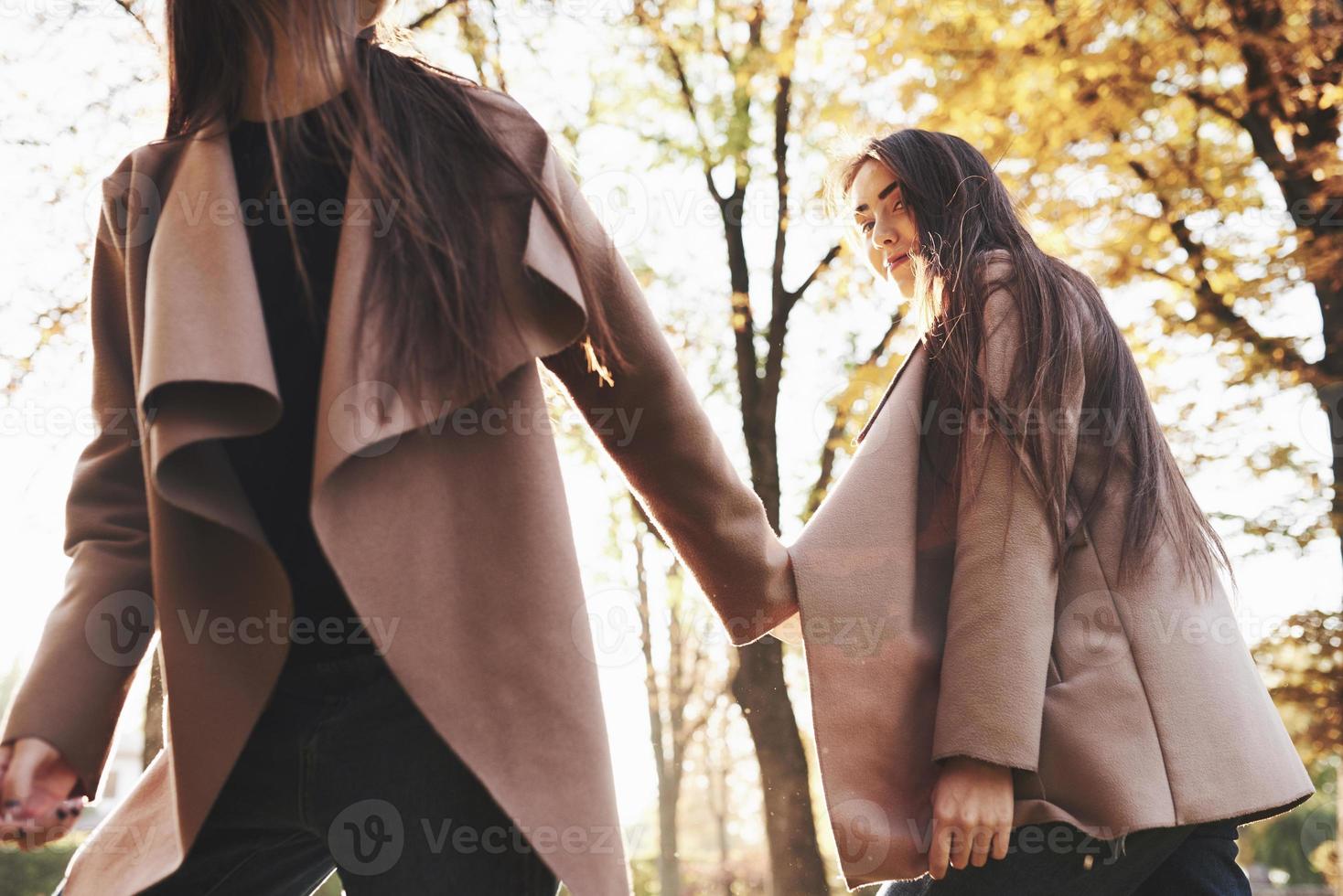 vista da parte inferior de jovens gêmeas morenas de mãos dadas e andando com um casaco casual no parque ensolarado de outono em fundo desfocado foto