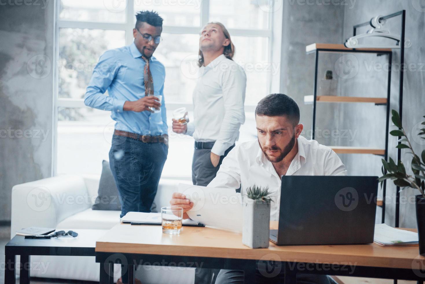 funcionários em segundo plano. jovem empresário se senta na cadeira em frente ao laptop e trabalha com relatórios foto