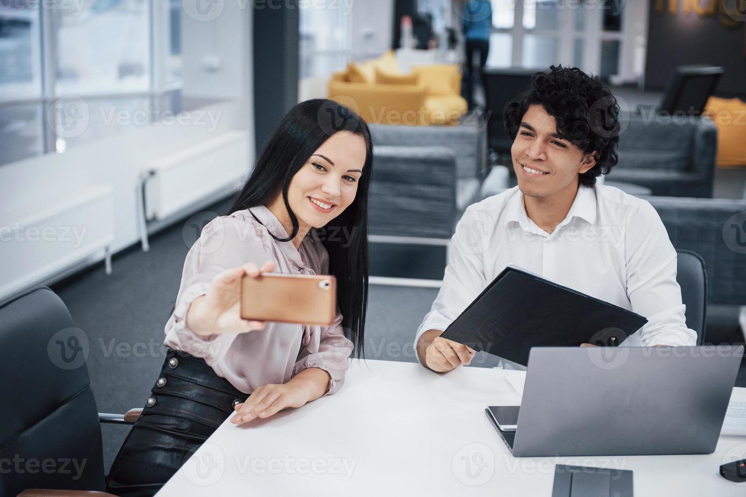 selfie de dois trabalhadores de escritório sorridentes com roupas oficiais sentados perto do laptop prateado na mesa foto