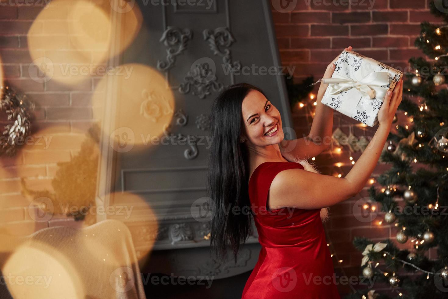 mulher feliz em um vestido vermelho segurando uma caixa branca de presente de Natal na sala com uma árvore de Natal e uma lareira atrás foto