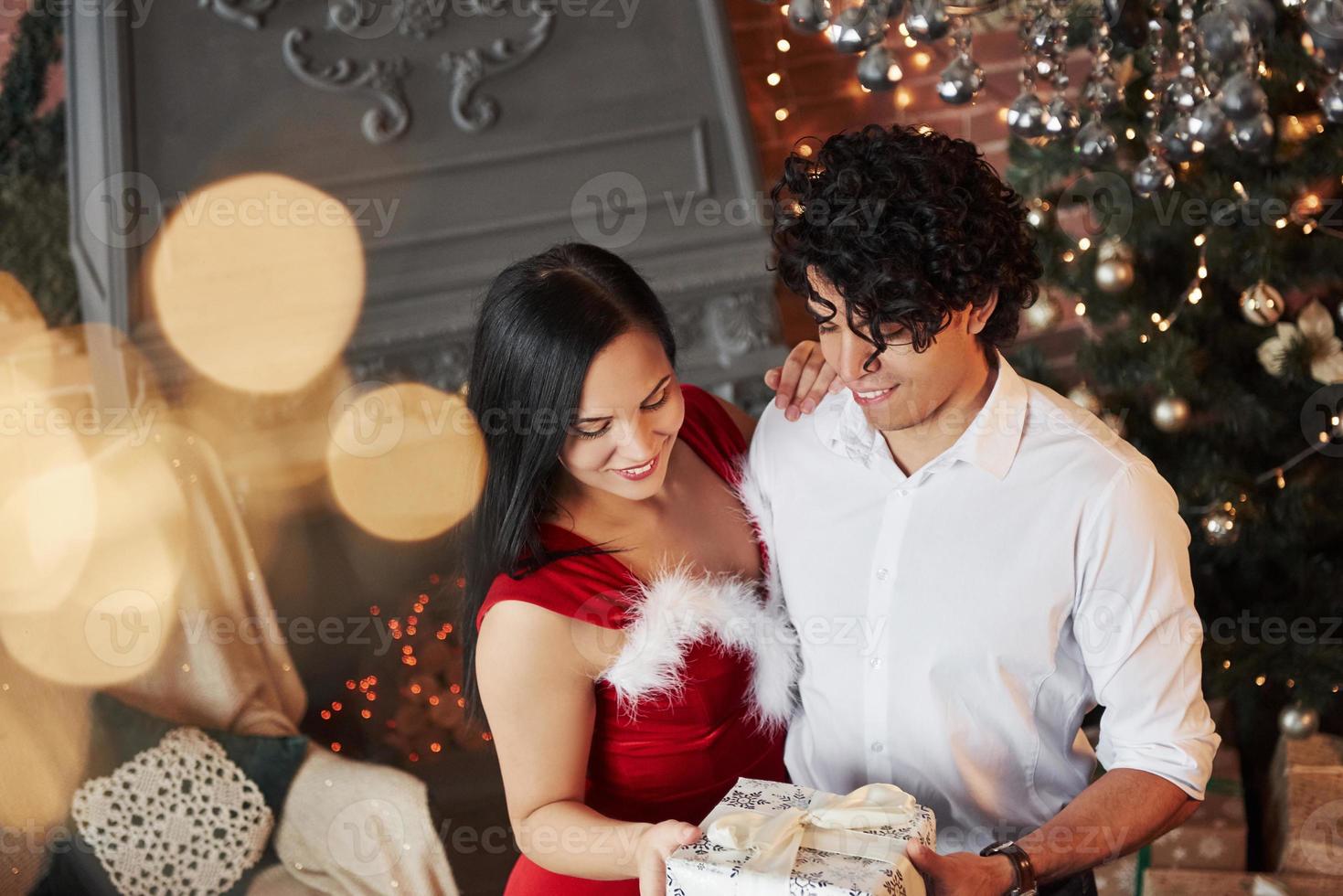 imaginando o que há dentro. lindo casal comemorando o ano novo na sala decorada com árvore de natal e lareira atrás foto