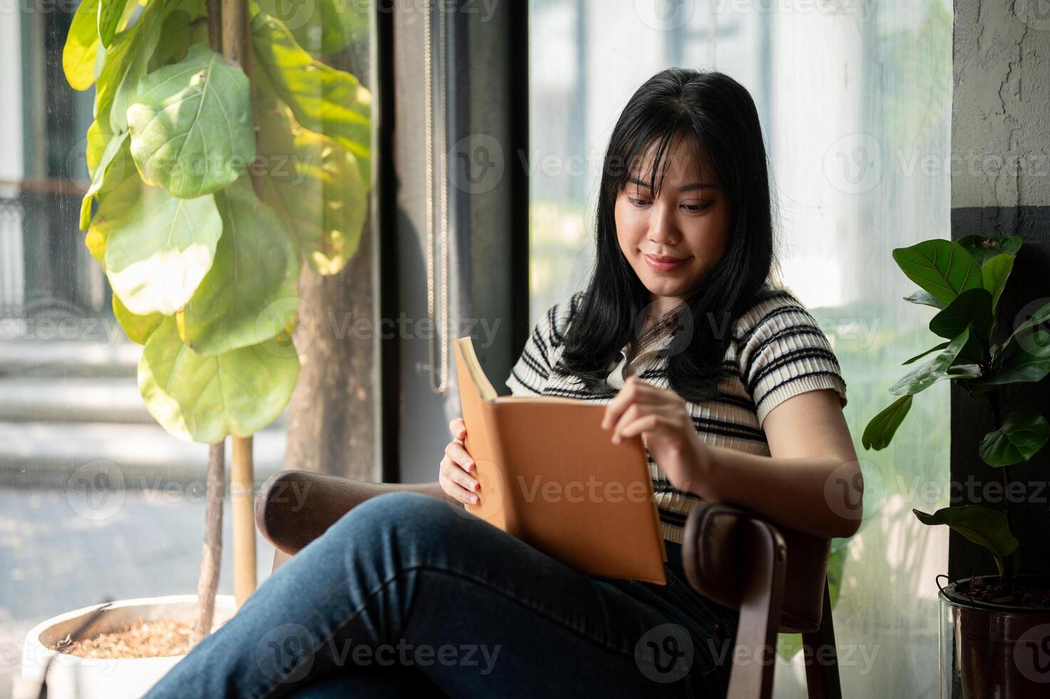 uma relaxado ásia mulher é lendo uma livro em a poltrona de a janela dentro uma lindo verde cafeteria. foto