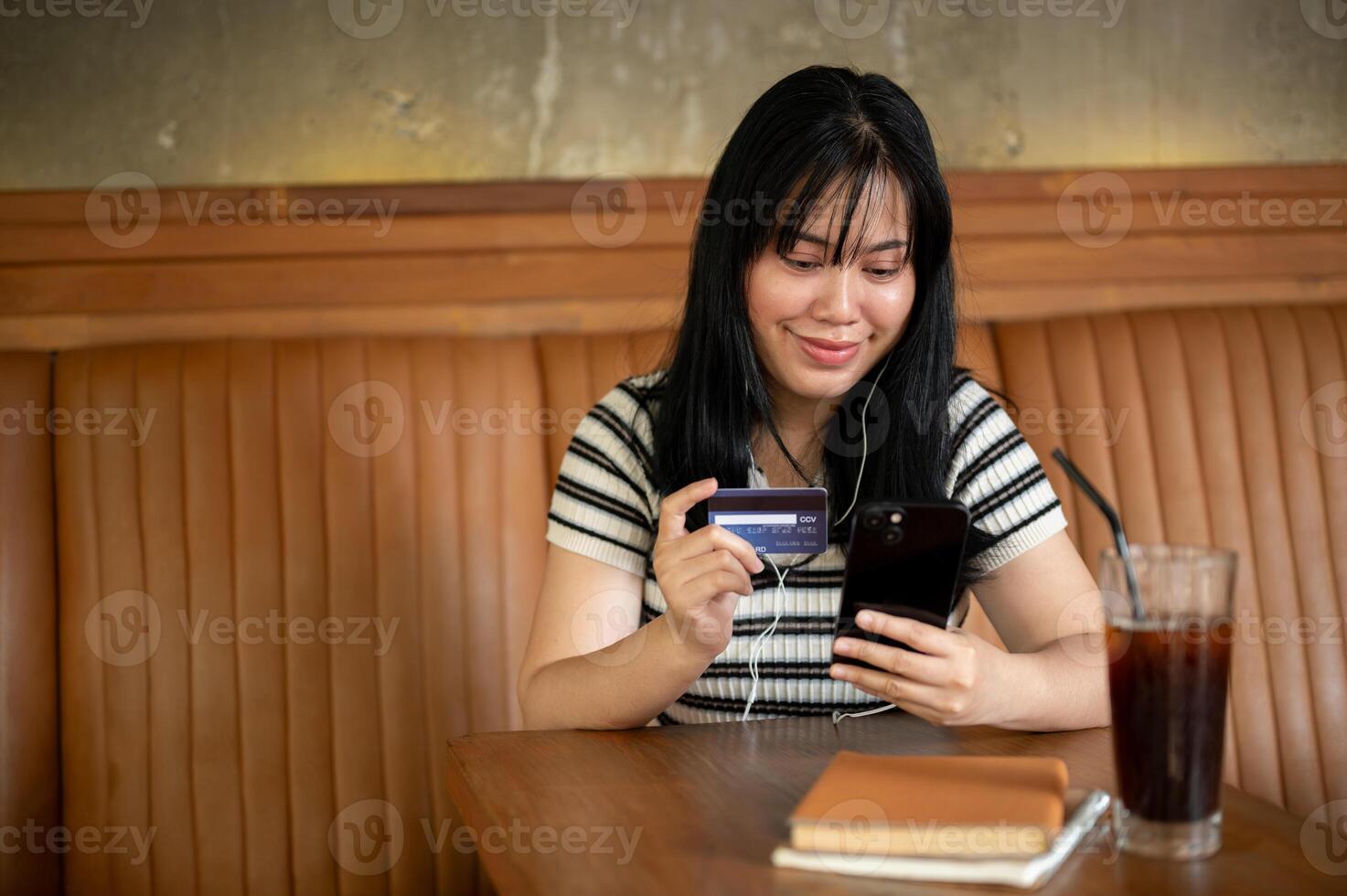uma mulher segurando uma Smartphone e uma crédito cartão às uma mesa dentro uma café comprar, pagando contas conectados foto
