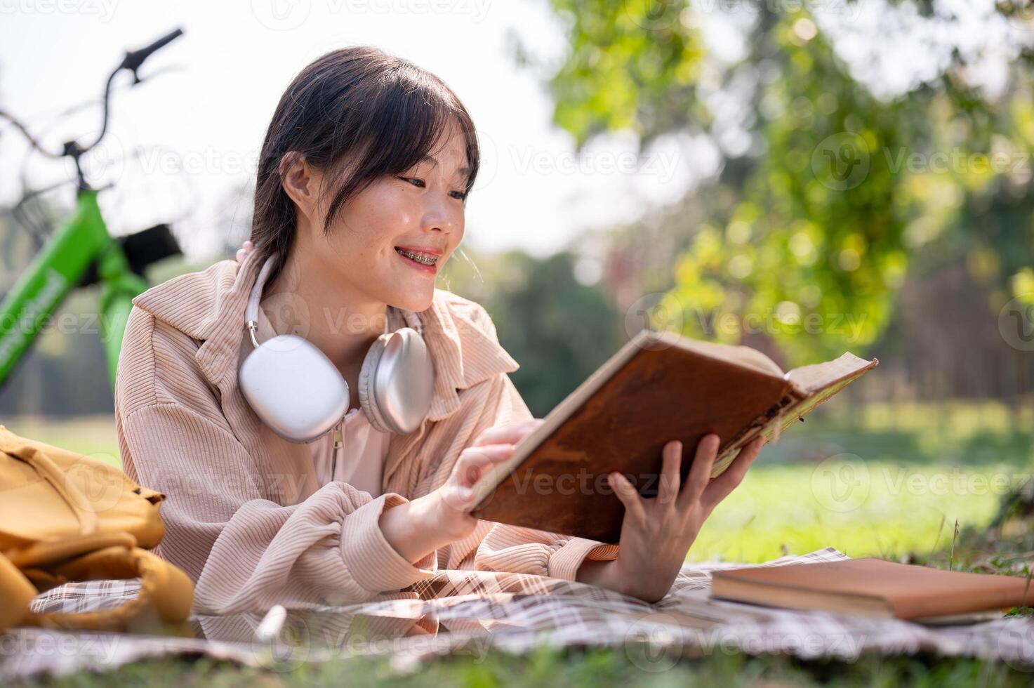 uma feliz ásia mulher é desfrutando lendo uma livro em uma piquenique pano, enquanto arrepiante dentro uma verde parque. foto