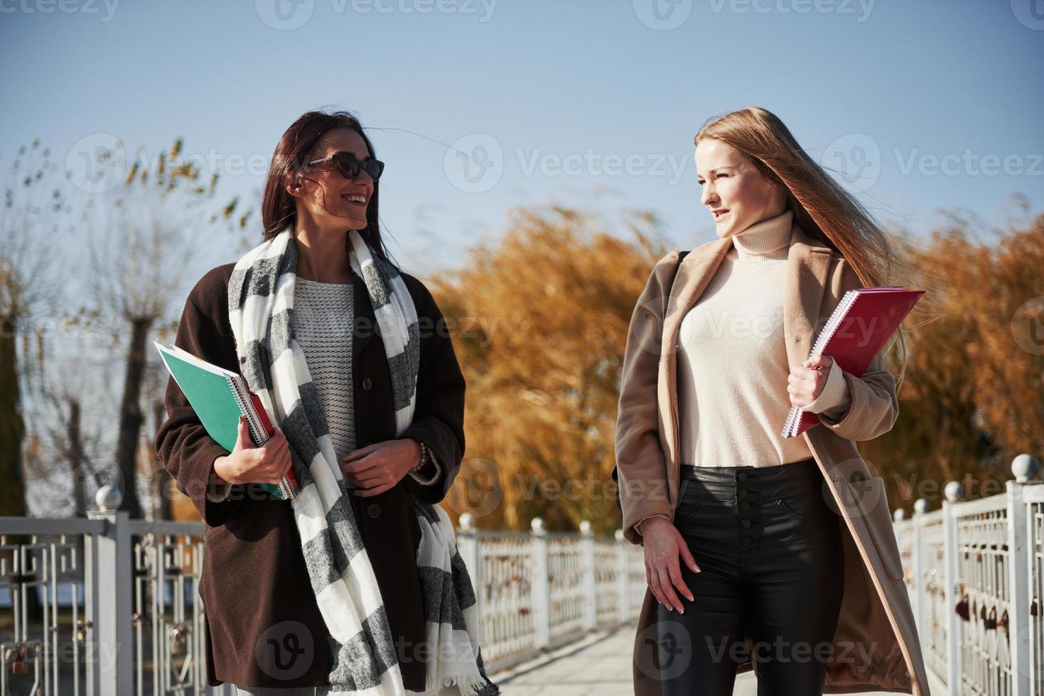 conversando. duas estudantes do sexo feminino caminham no parque na ponte segurando blocos de notas foto