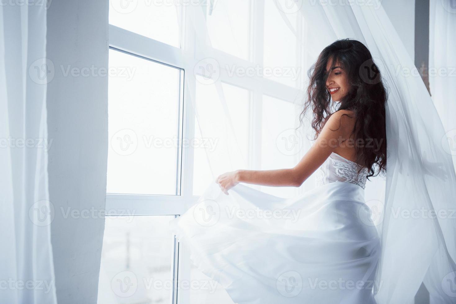 atrás das cortinas. Mulher bonita em um vestido branco fica em um quarto branco com a luz do dia entrando pelas janelas foto