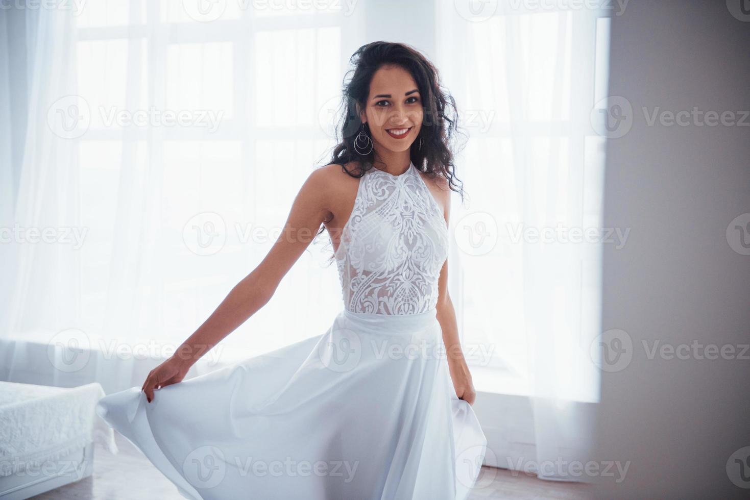 roupa clássica para danças e festas luxuosas. Mulher bonita em um vestido branco fica em um quarto branco com a luz do dia entrando pelas janelas foto