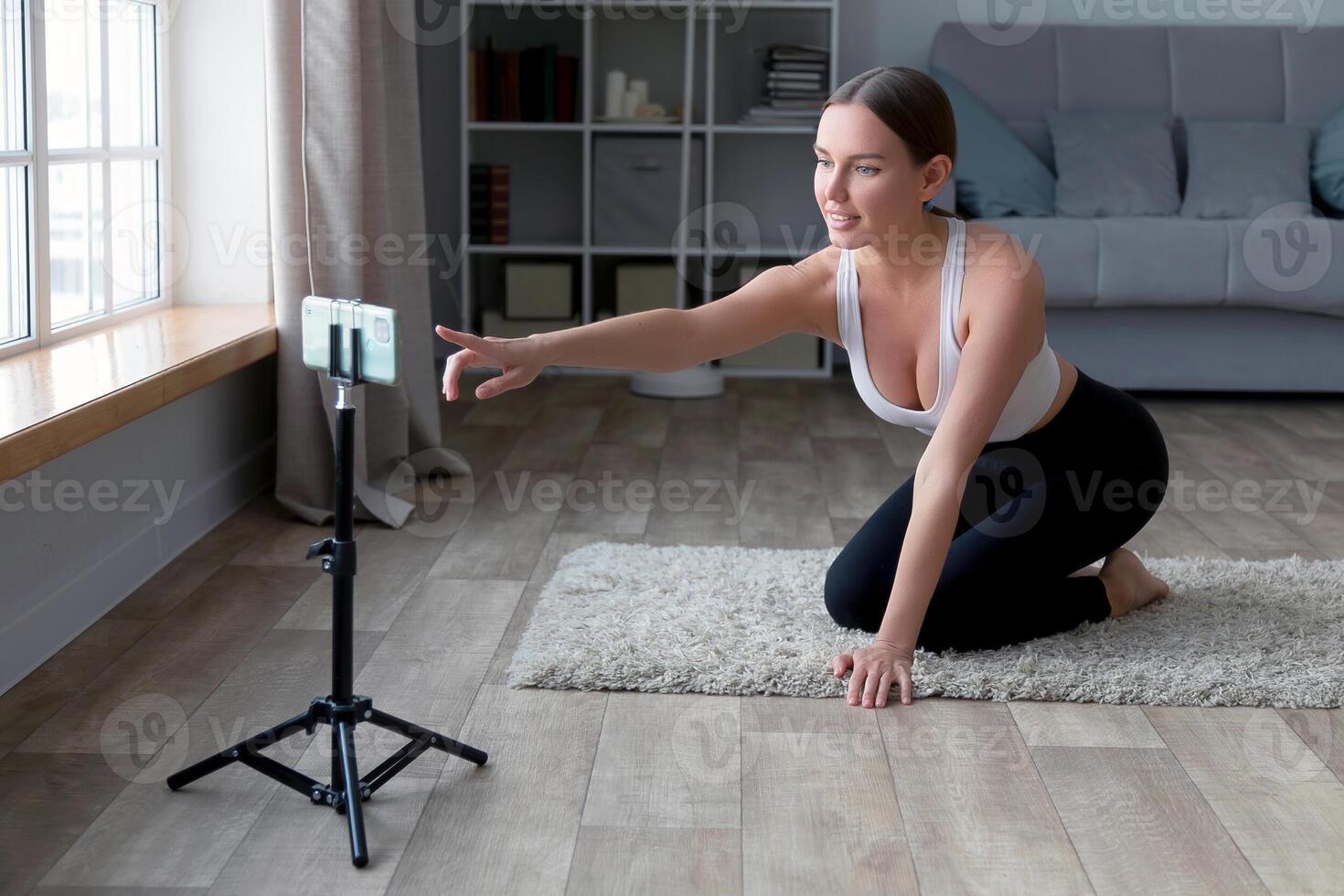 mulher sentado em a chão dentro ioga calça gravação a conectados ginástica com dela telefone e tripé, sorridente às a Câmera dentro uma vivo quarto configuração foto