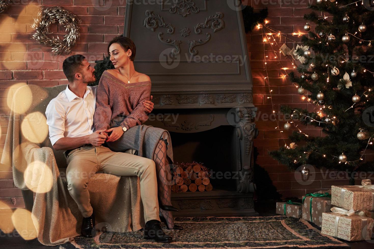 lindo casal jovem na cadeira se abraçando em um quarto de luxo decorado com árvore de natal e guirlandas foto