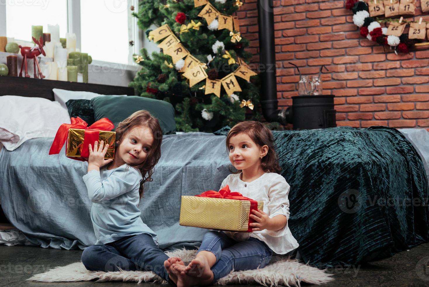 cada um deles tem um bom presente. férias de natal com presentes para essas duas crianças sentadas dentro de casa no belo quarto perto da cama foto