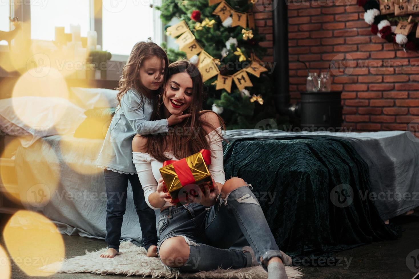 olha o que eu tenho para você. mãe e filha sentadas em um quarto com decoração festiva segurando uma caixa de presente foto