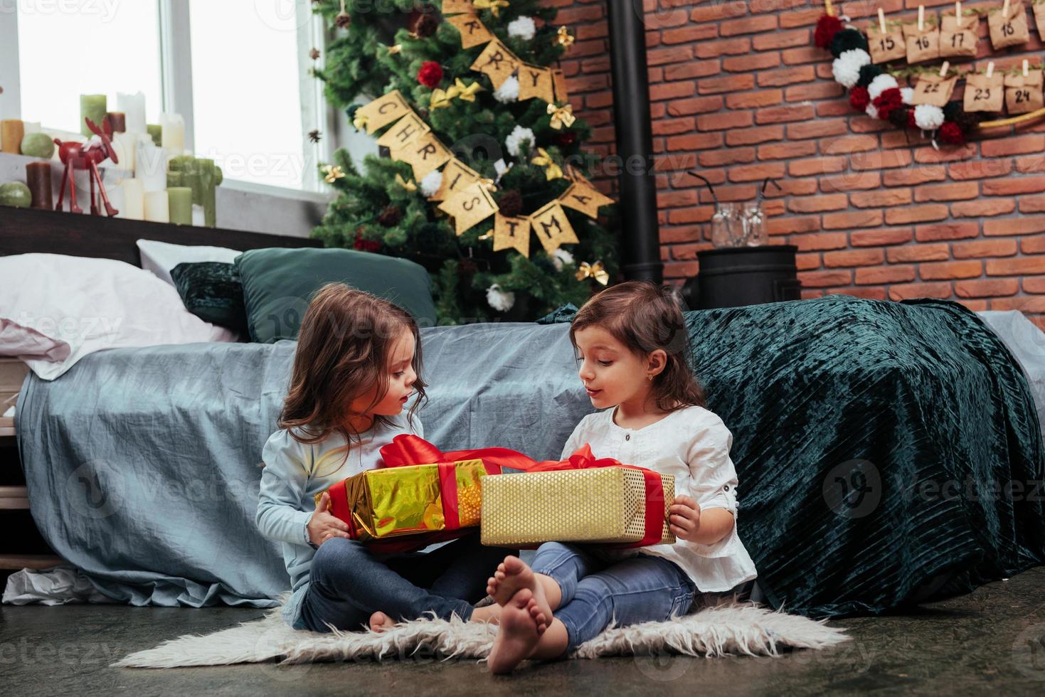 falando enquanto segura caixas coloridas. férias de natal com presentes para essas duas crianças sentadas dentro de casa no belo quarto perto da cama foto