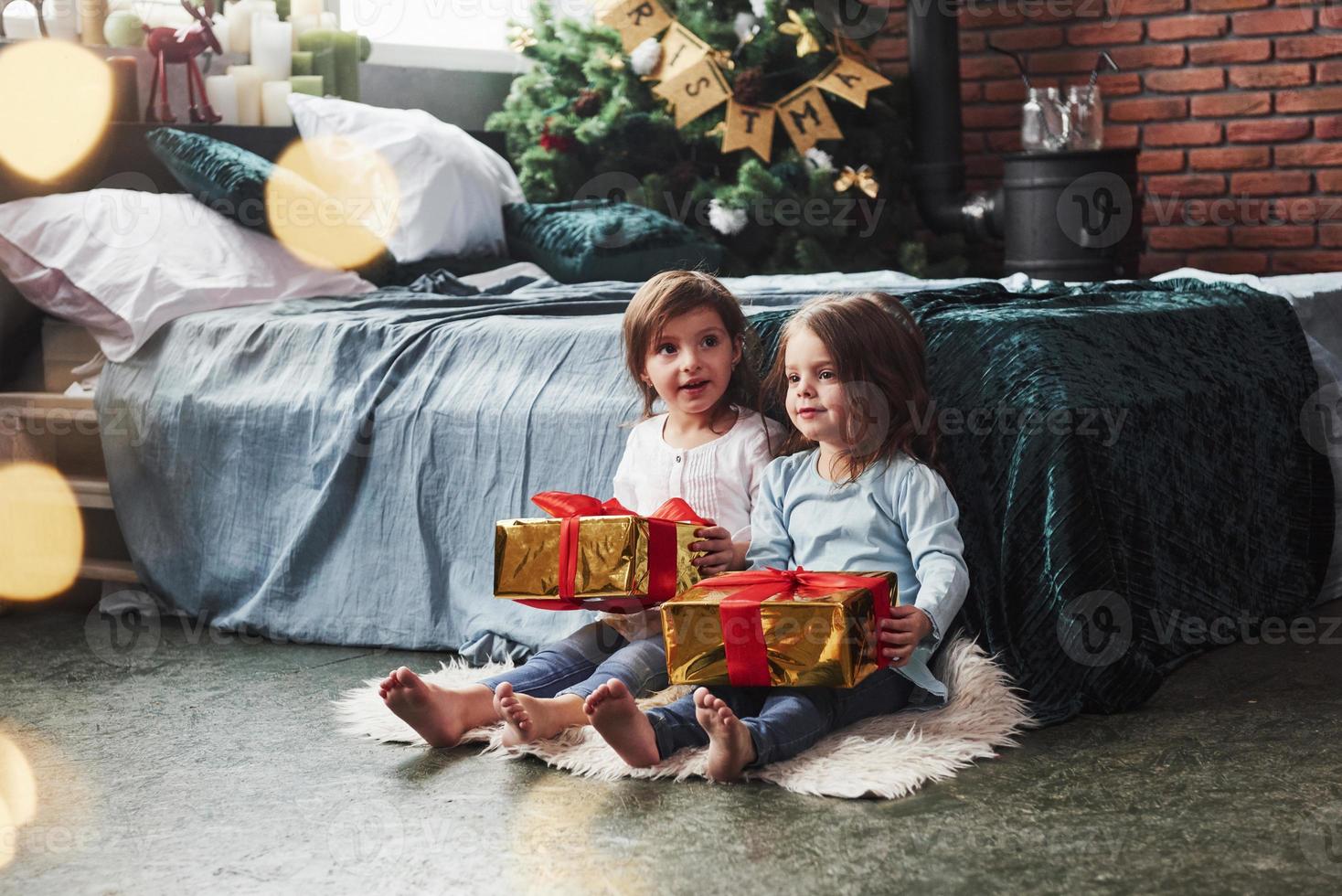 hoje alguém tem seu presente. férias de natal com presentes para essas duas crianças sentadas dentro de casa no belo quarto perto da cama foto