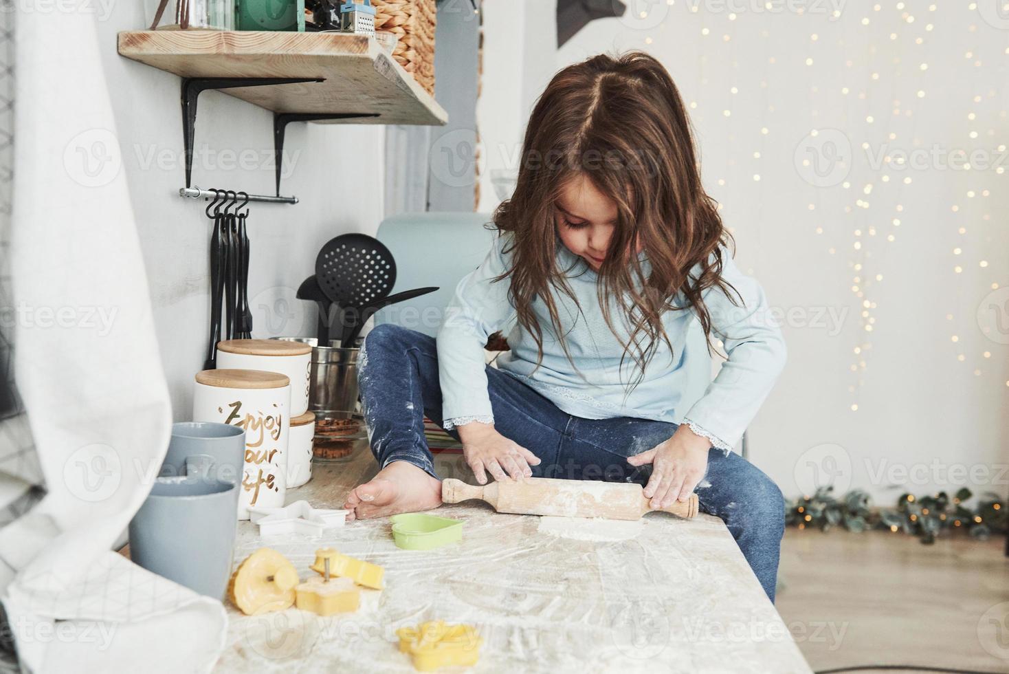 guirlandas e decorações do feriado no fundo. foto de uma linda garotinha sentada na mesa da cozinha brincando com farinha