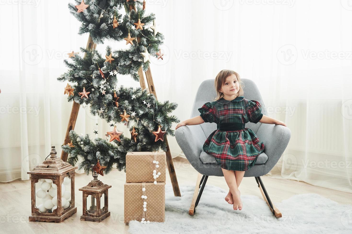 bela sala branca. concepção de natal e feriados. menina bonitinha está sentada na cadeira perto da escada decorada com estrelas e caixas de presente no chão foto