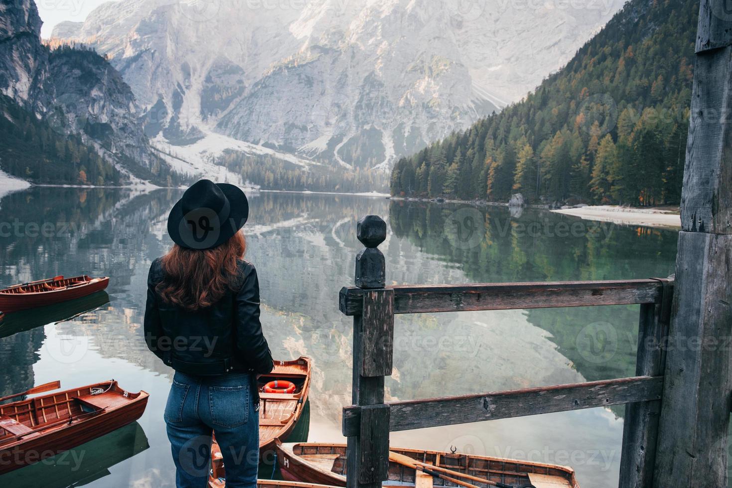 madeiras e rochas pendentes. mulher de chapéu preto apreciando a paisagem montanhosa majestosa perto do lago com barcos foto