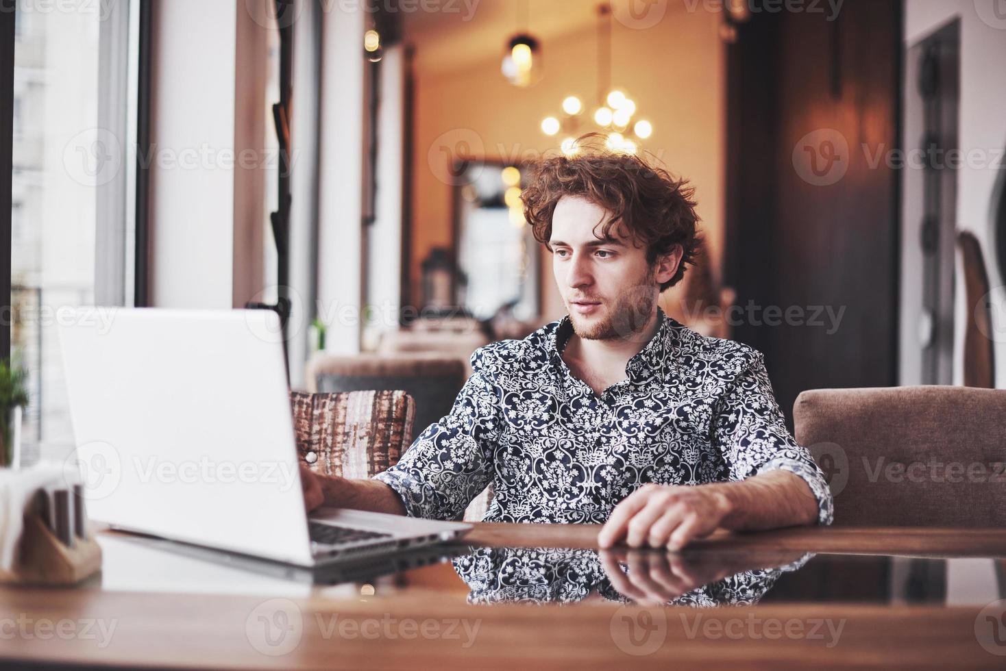 jovem bonito sentado no escritório com uma xícara de café e trabalhando em um projeto conectado com tecnologias cibernéticas modernas. empresário com notebook tentando cumprir prazos na esfera do marketing digital foto
