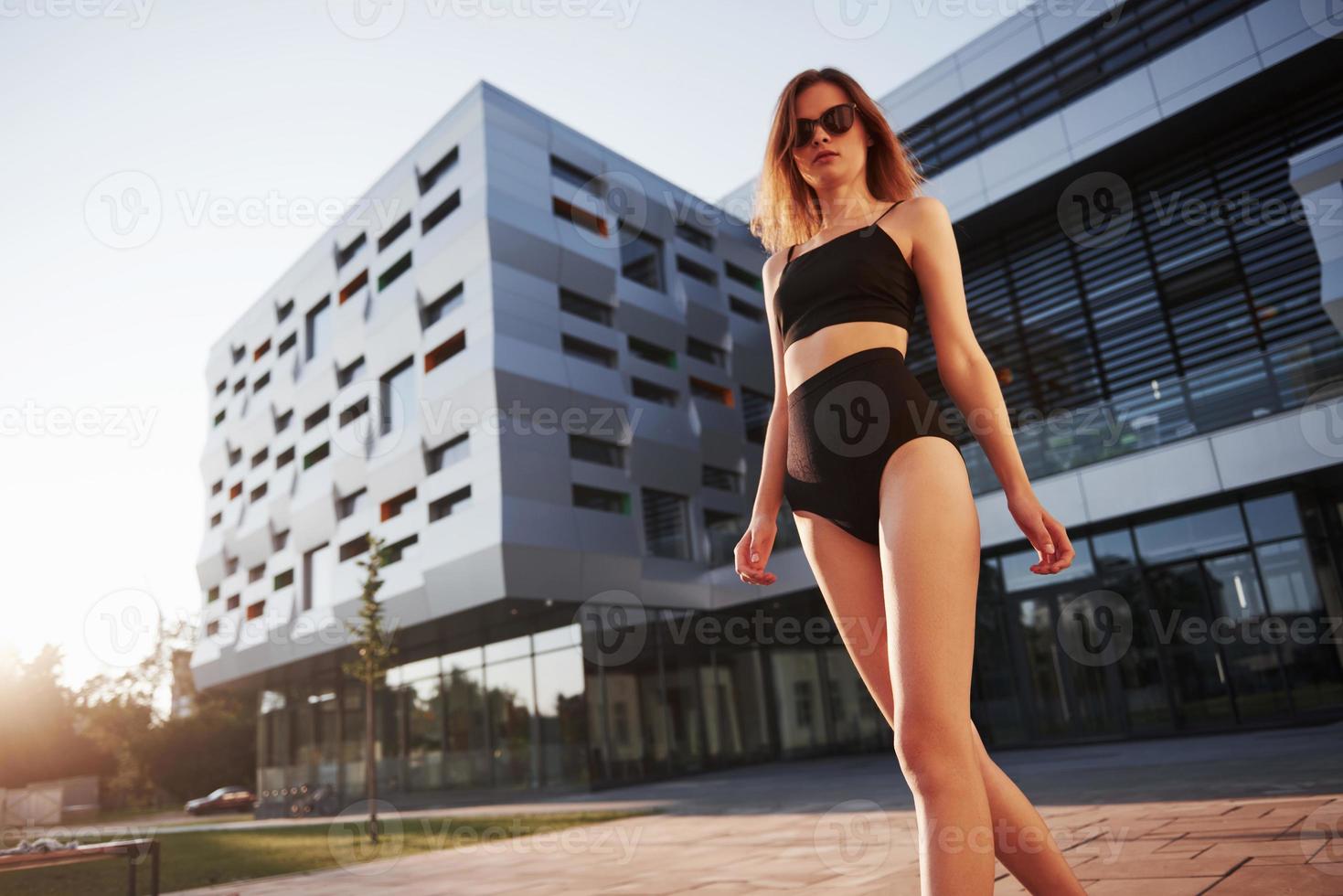 mulher jovem sexy com óculos escuros e maiô preto está de pé no fundo dos edifícios da cidade durante o pôr do sol foto