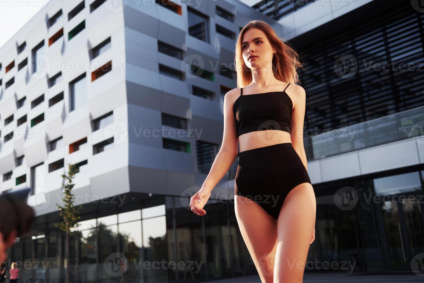 mulher jovem sexy com óculos escuros e maiô preto está de pé no fundo dos edifícios da cidade durante o pôr do sol foto