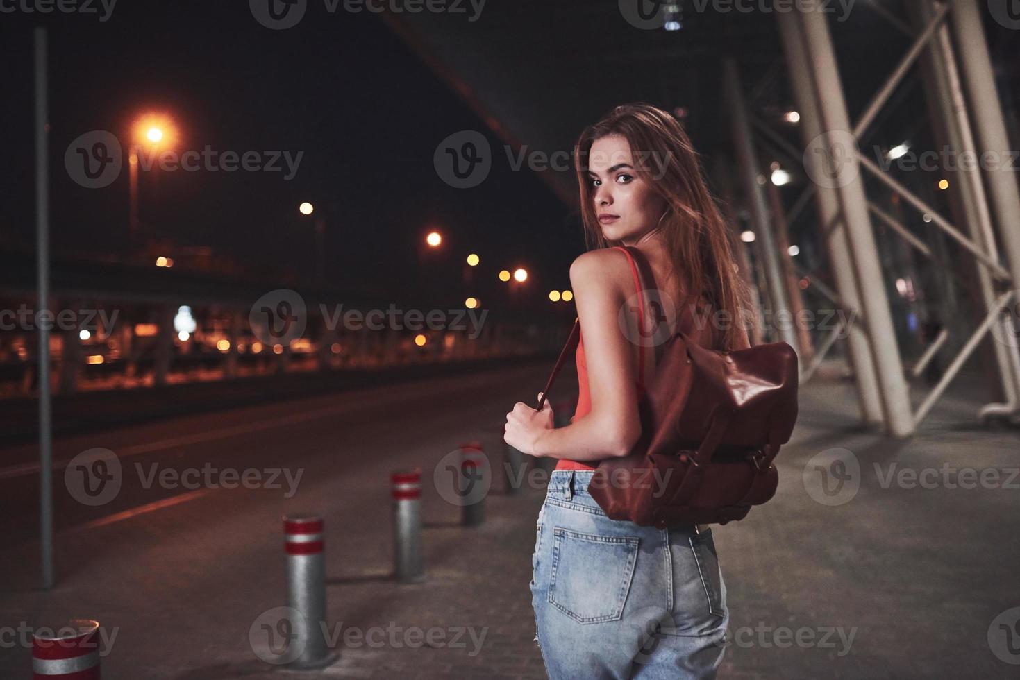 uma linda jovem com uma mochila atrás dos ombros está na rua perto de um aeroporto ou estação ferroviária em uma noite quente de verão. ela acabou de chegar e espera por um táxi ou seus amigos foto