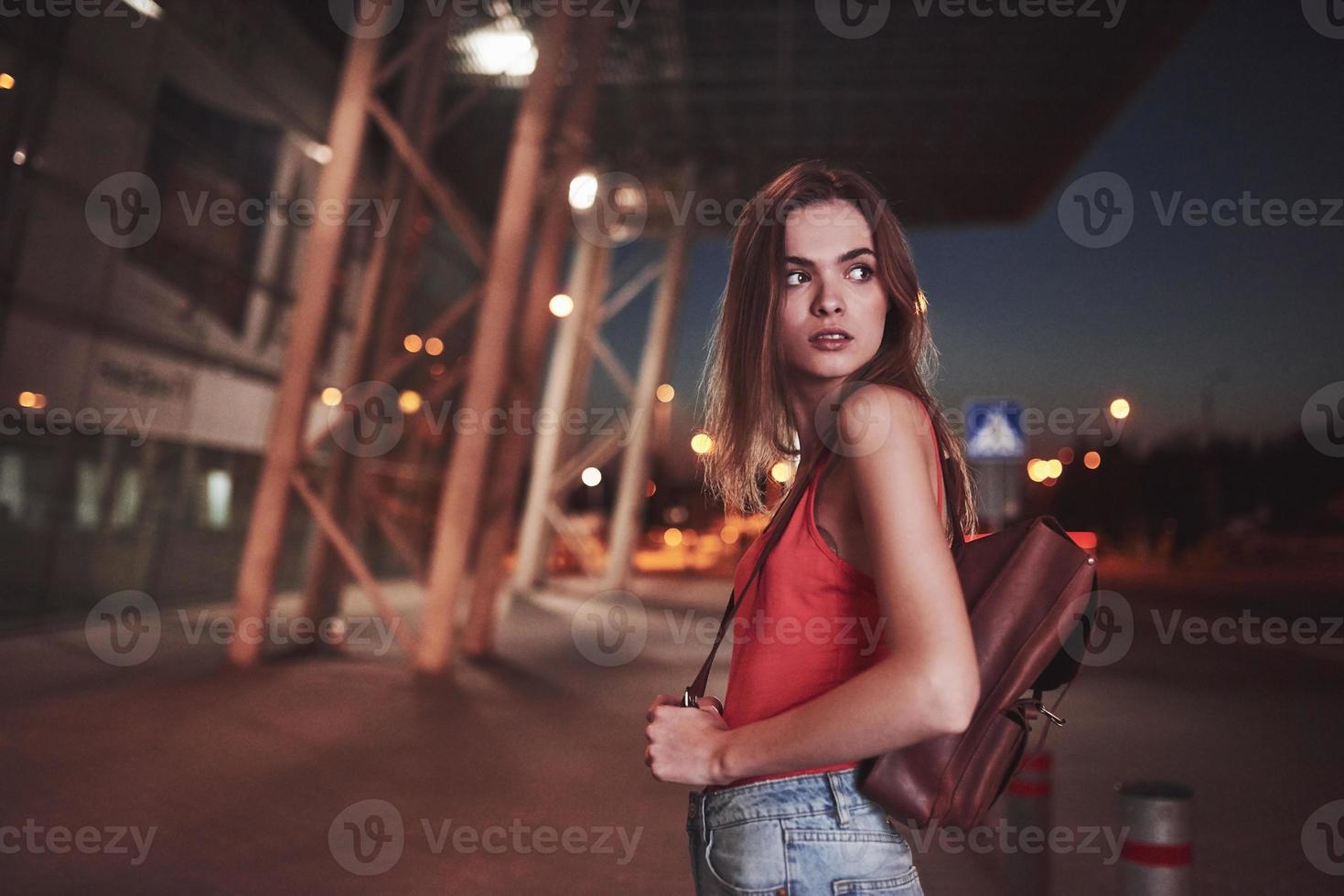 uma linda jovem com uma mochila atrás dos ombros está na rua perto de um aeroporto ou estação ferroviária em uma noite quente de verão. ela acabou de chegar e espera por um táxi ou seus amigos foto