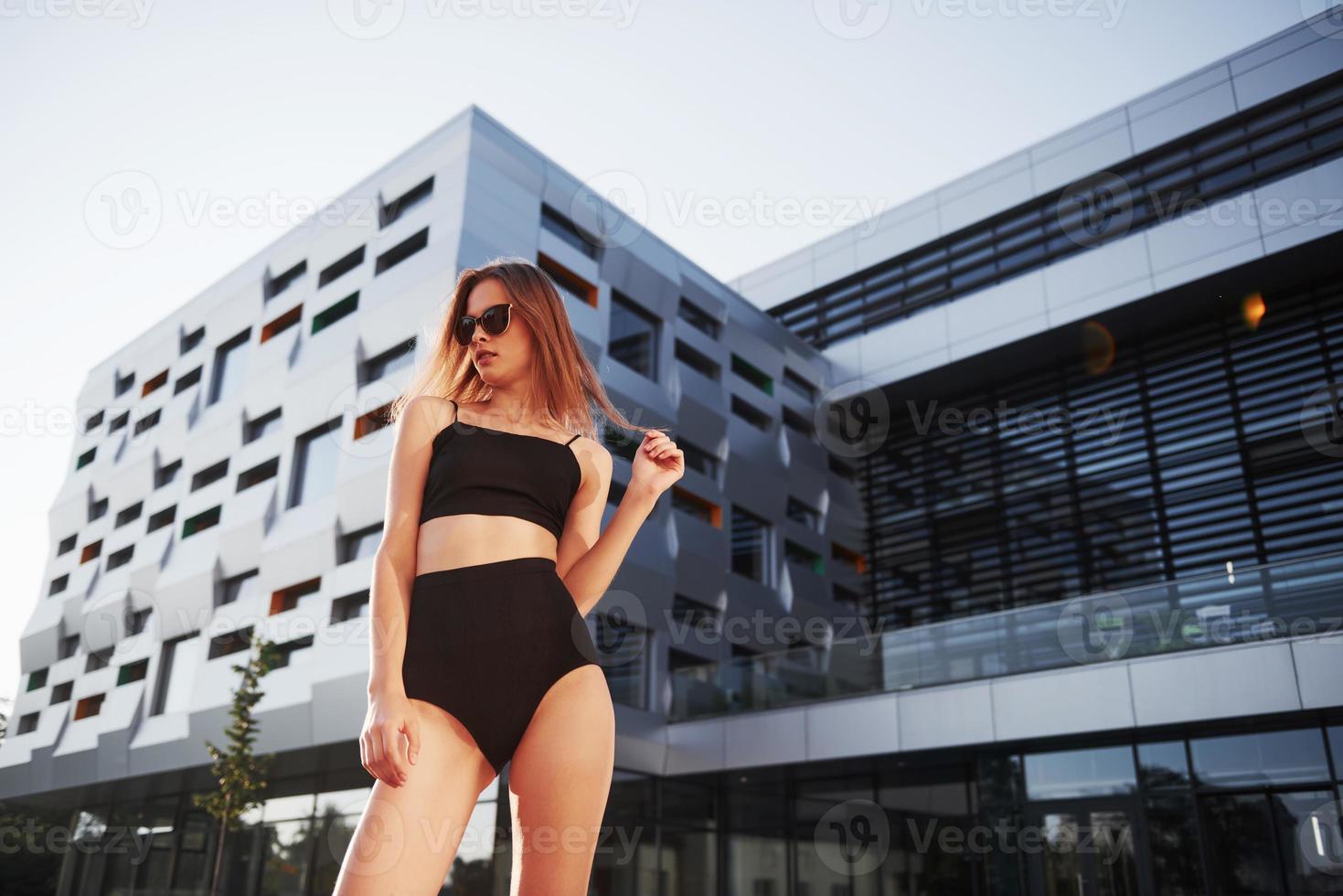 mulher jovem sexy com óculos escuros e maiô preto está de pé no fundo dos edifícios da cidade durante o pôr do sol foto