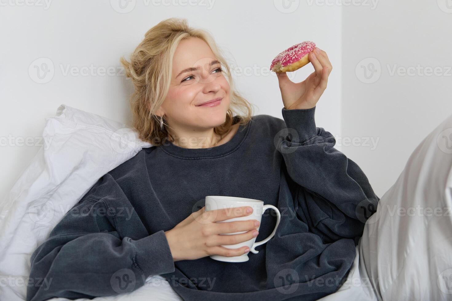 fechar acima retrato do lindo sorridente, loiro mulher, deitado dentro cama, bebidas chá e come rosquinha em uma preguiçoso final de semana ou folga foto