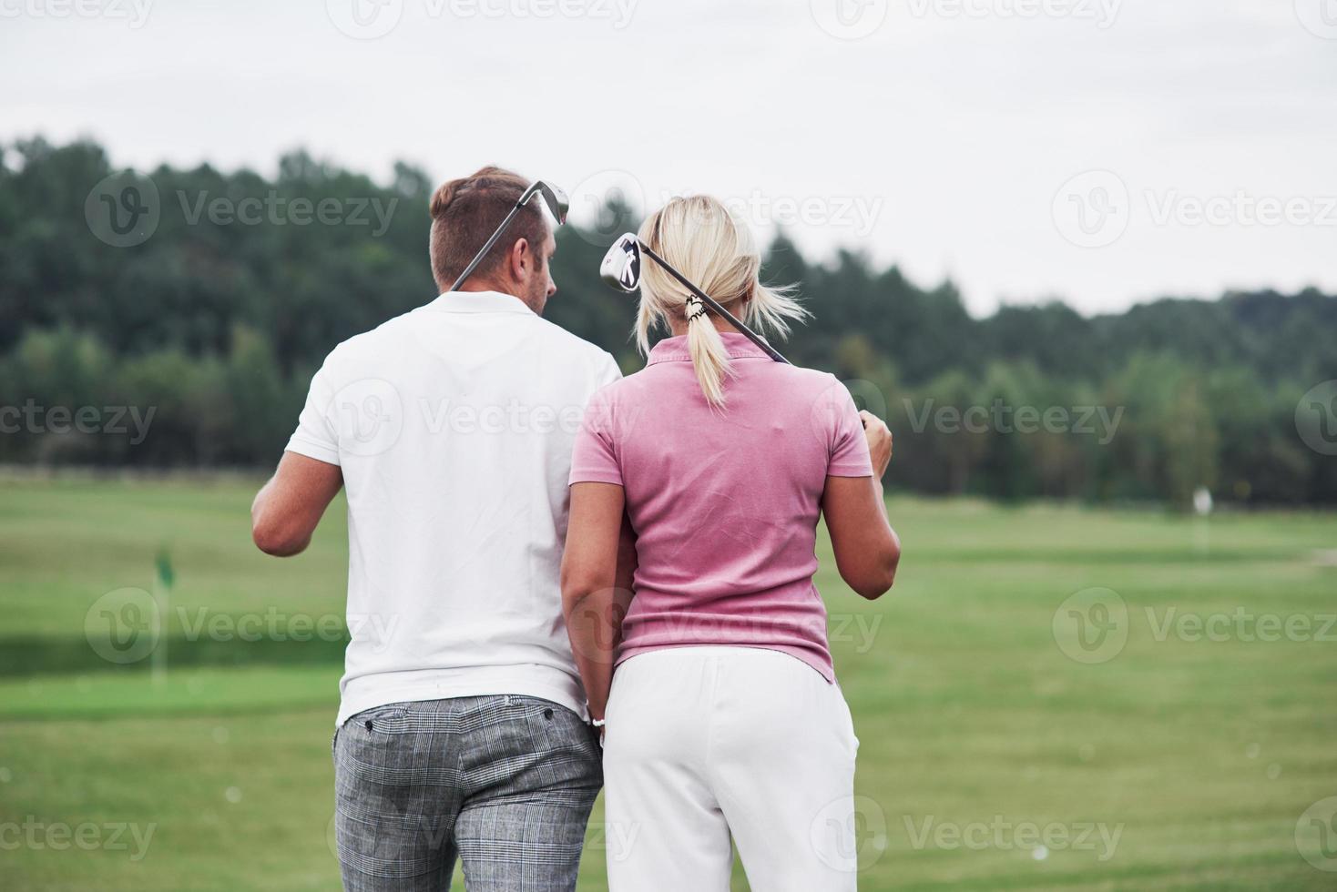 vista traseira. casal de jogadores de golfe com tacos nas mãos caminhando no gramado foto