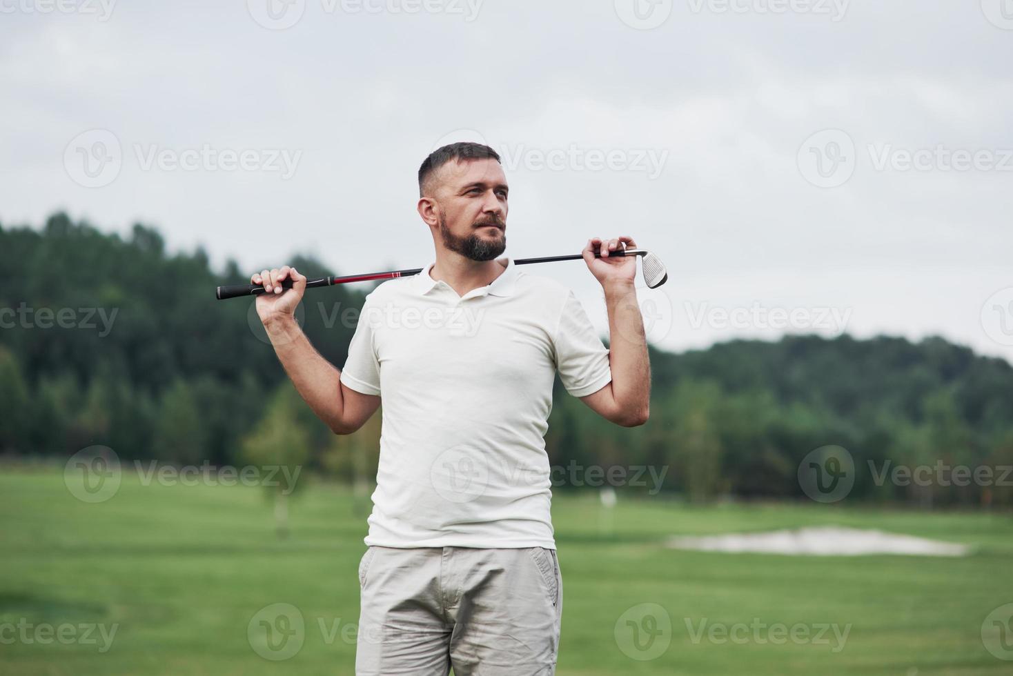 olhando para longe. retrato do jogador de golfe em pé no gramado e taco na mão. bosques no fundo foto