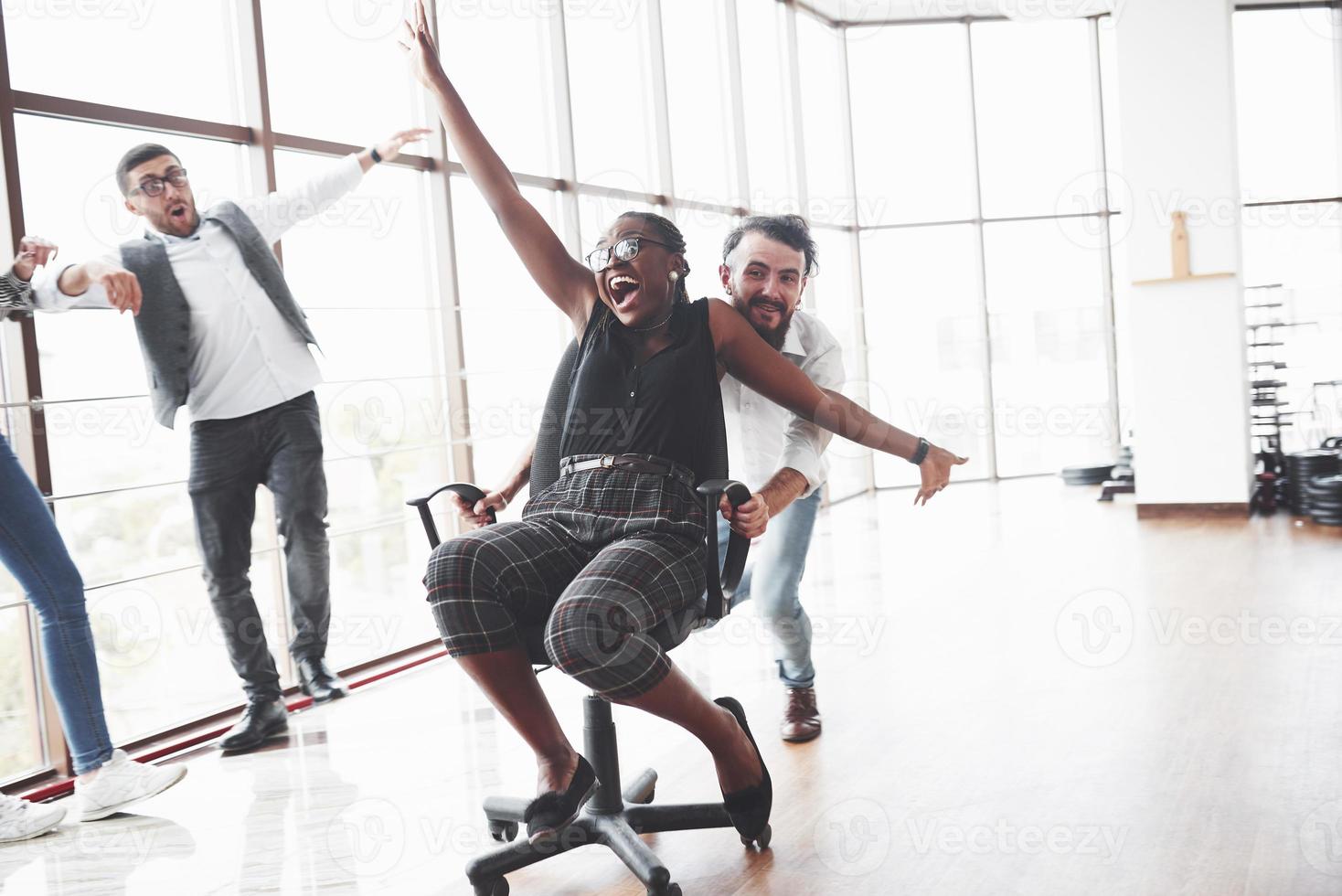 jovens se divertindo na espaçosa sala de escritório montando o afro-americano oman na cadeira foto