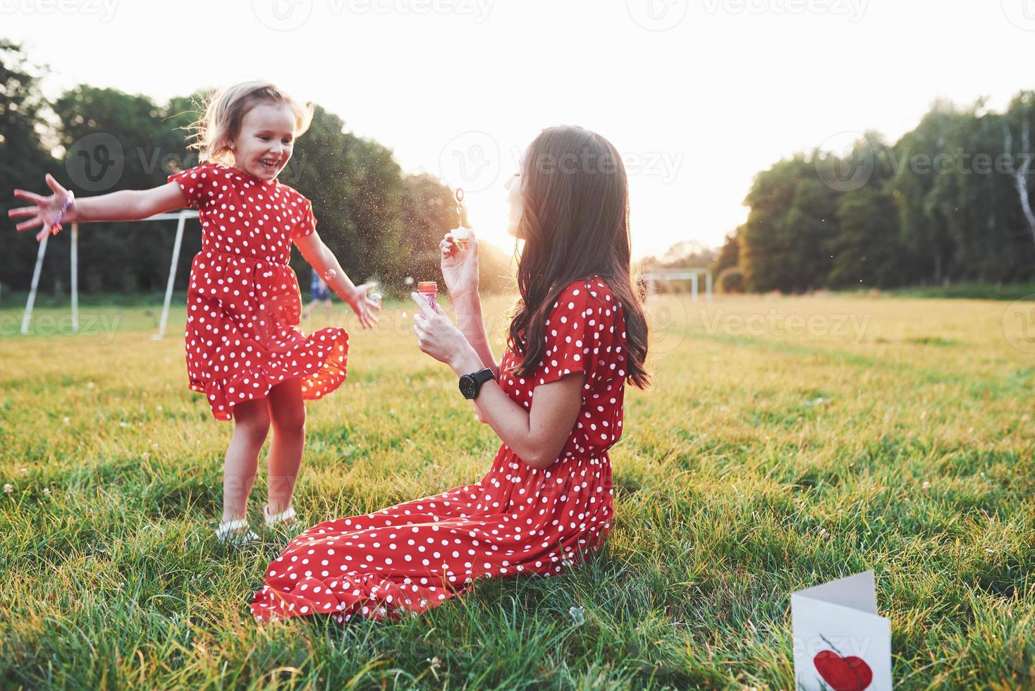 me dê mais. menina com a filha se divertindo com bolhas do lado de fora, sentada na grama foto