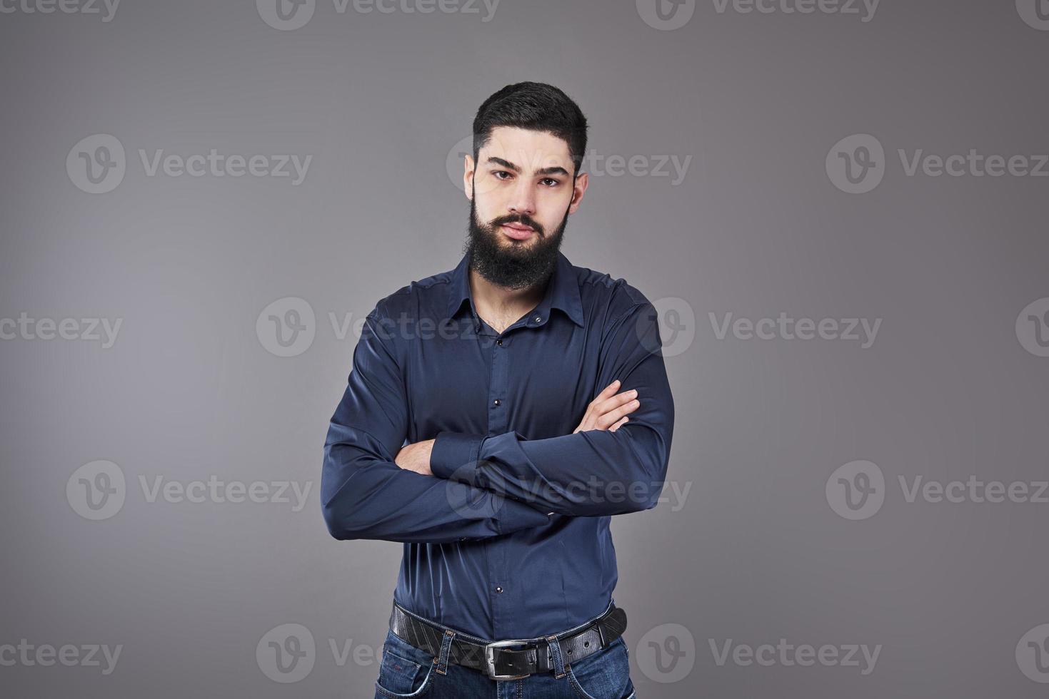 jovem bonito encostado na parede cinza com os braços cruzados. um jovem sério com barba olha para a câmera foto