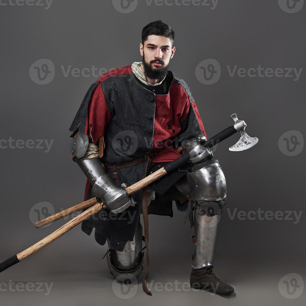 cavaleiro medieval em fundo cinza. retrato de guerreiro brutal com rosto sujo com cota de malha, roupas vermelhas e pretas e machado de batalha foto