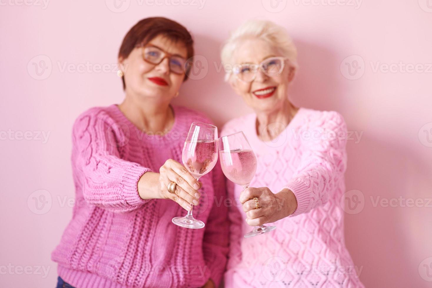 duas elegantes mulheres sêniors em suéteres cor de rosa bebendo vinho rosé na cozinha moderna fofocando. amizade, conversa, fofoca, evento, relacionamentos, notícias, conceito de família foto