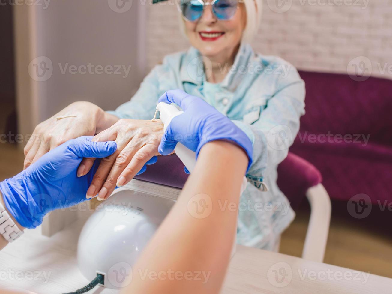 Mestre de manicure com luvas azuis cremosando as mãos de uma mulher idosa elegante em óculos de sol azuis e jaqueta jeans sentada no salão de manicure foto