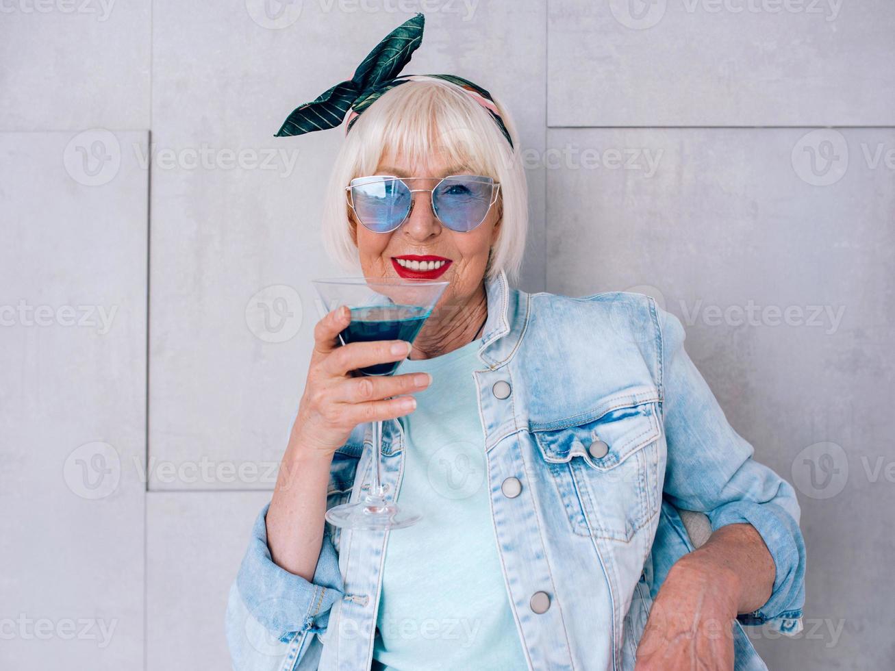 mulher elegante sênior com cabelos grisalhos e óculos azuis e jaqueta jeans, segurando um copo com coquetel azul. álcool, relaxar, férias, conceito de aposentadoria foto
