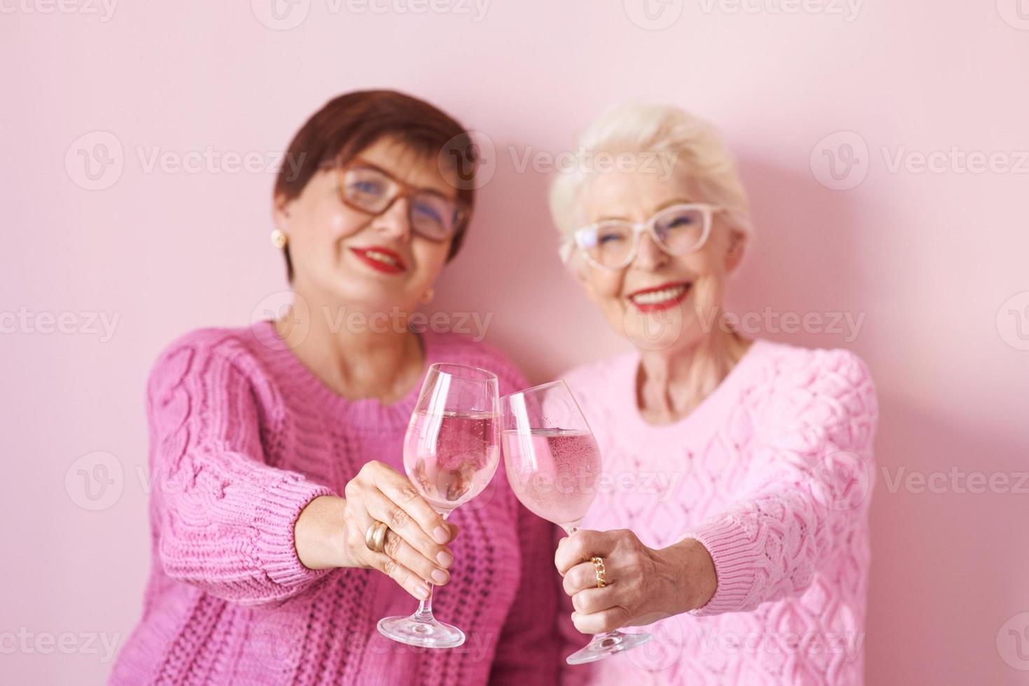 duas elegantes mulheres sêniors em suéteres cor de rosa bebendo vinho rosé na cozinha moderna fofocando. amizade, conversa, fofoca, evento, relacionamentos, notícias, conceito de família foto